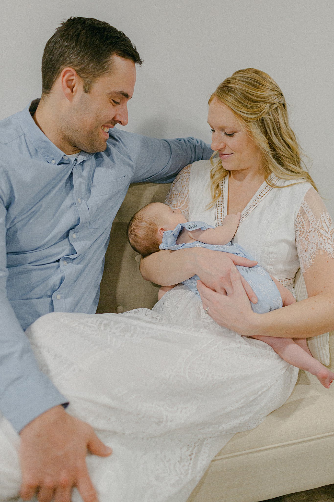 Mom in white dress, holding baby girl in blue romper, with Dad smiling down at her at a Newborn Session in Green Bay